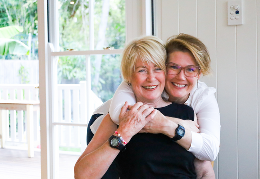 Blonde woman wearing a black t-shirt being hugged from behind by a darker blonde woman wearing glasses and a long sleeve white shirt