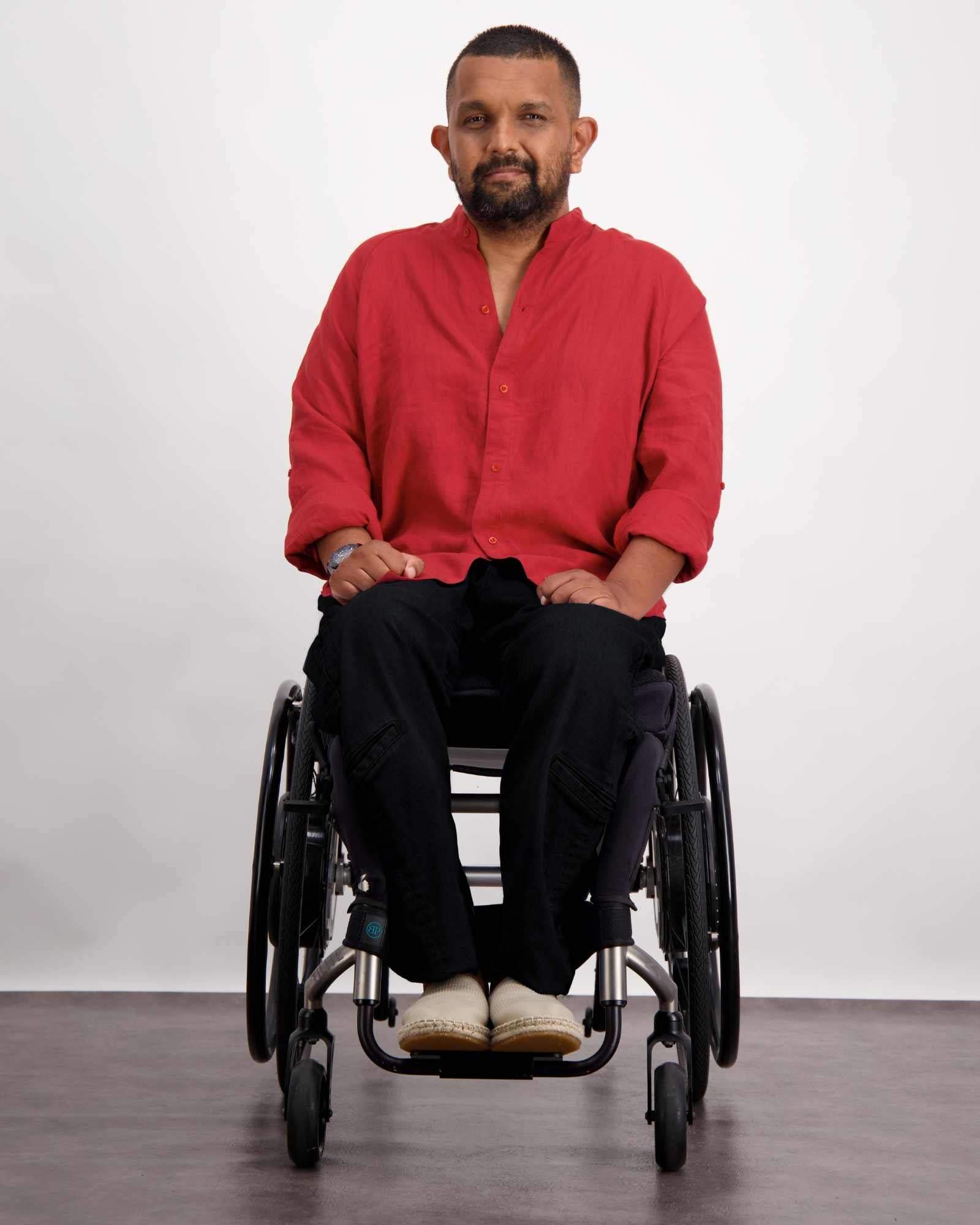 A person in a wheelchair wearing the Stephen Seated Jeans in black, paired with a vibrant red button-up shirt with rolled sleeves. The adaptive jeans feature a comfortable seated design. The model is photographed against a light background, showcasing the casual yet stylish ensemble.