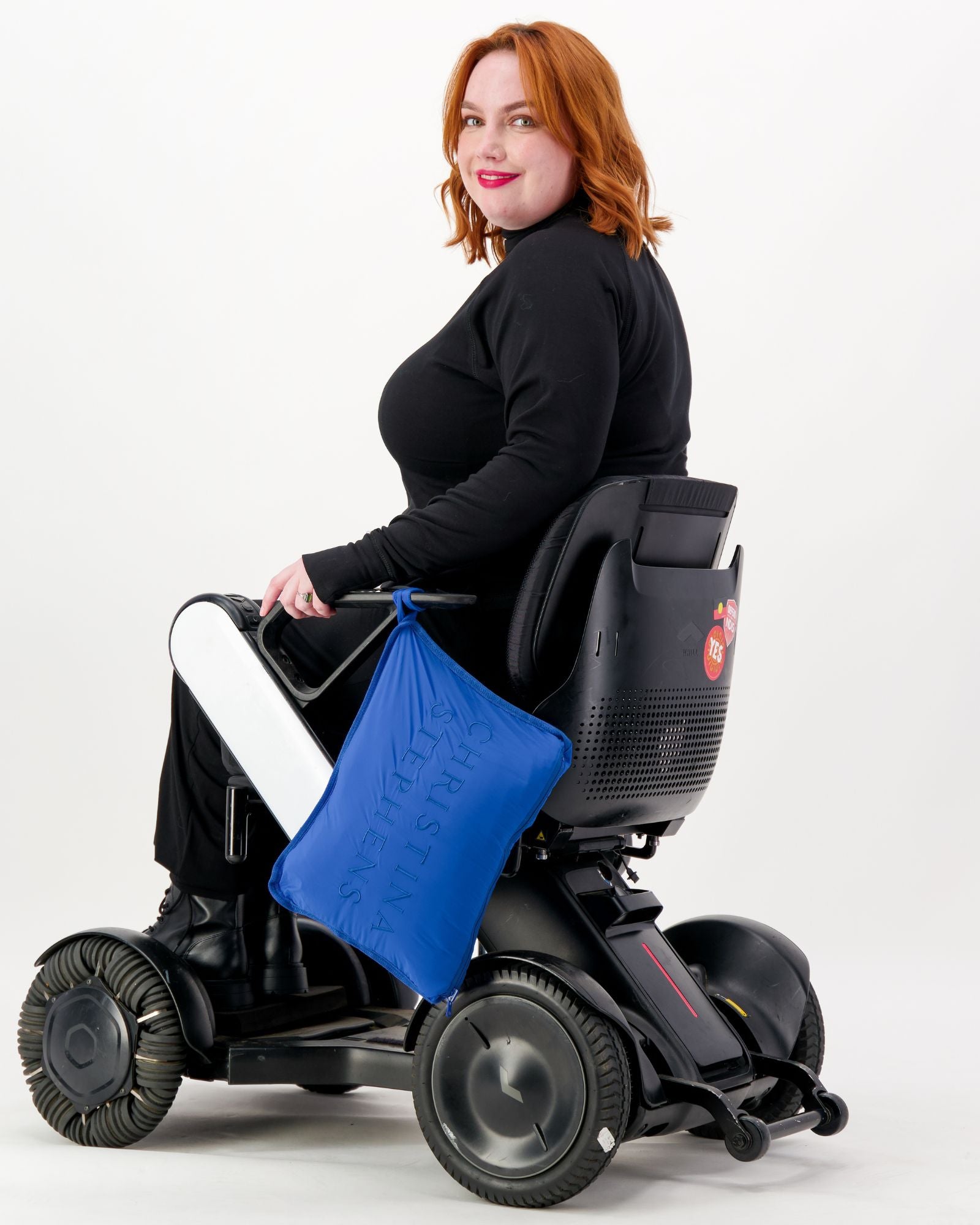 The Maggie Thermal Top shown on a model seated in a power wheelchair, featuring a classic black design with long sleeves. A bright blue accessory adds a pop of color against the sleek black garment, demonstrating its adaptable style for wheelchair users.