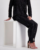 Suzie Q Seated Pants Sequins shown on a white pedestal, featuring sleek black sequined fabric with a comfortable adaptive design. The pants showcase elegant sparkle detail from hip to ankle, with a relaxed fit and specialized accommodations for medical devices.