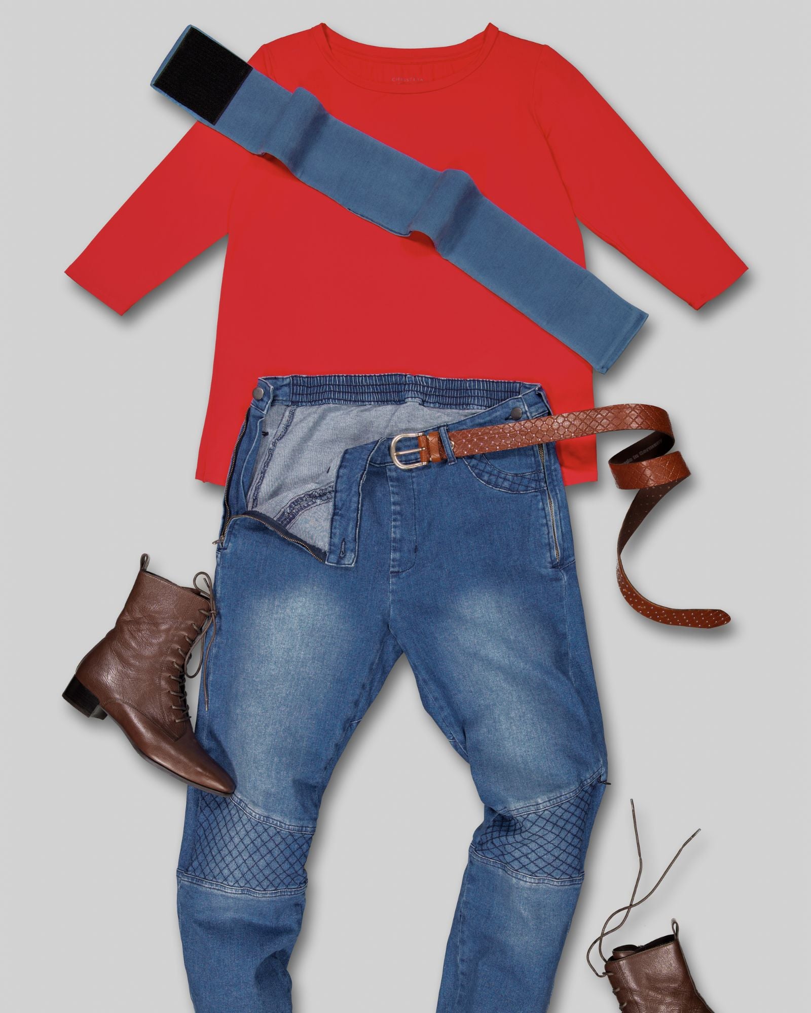 Wheelchair Leg Strap shown with red sweater, adaptive denim jeans, brown leather belt and boots in a flatlay composition. The denim blue strap features a secure black fastening mechanism for comfortable wheelchair positioning.