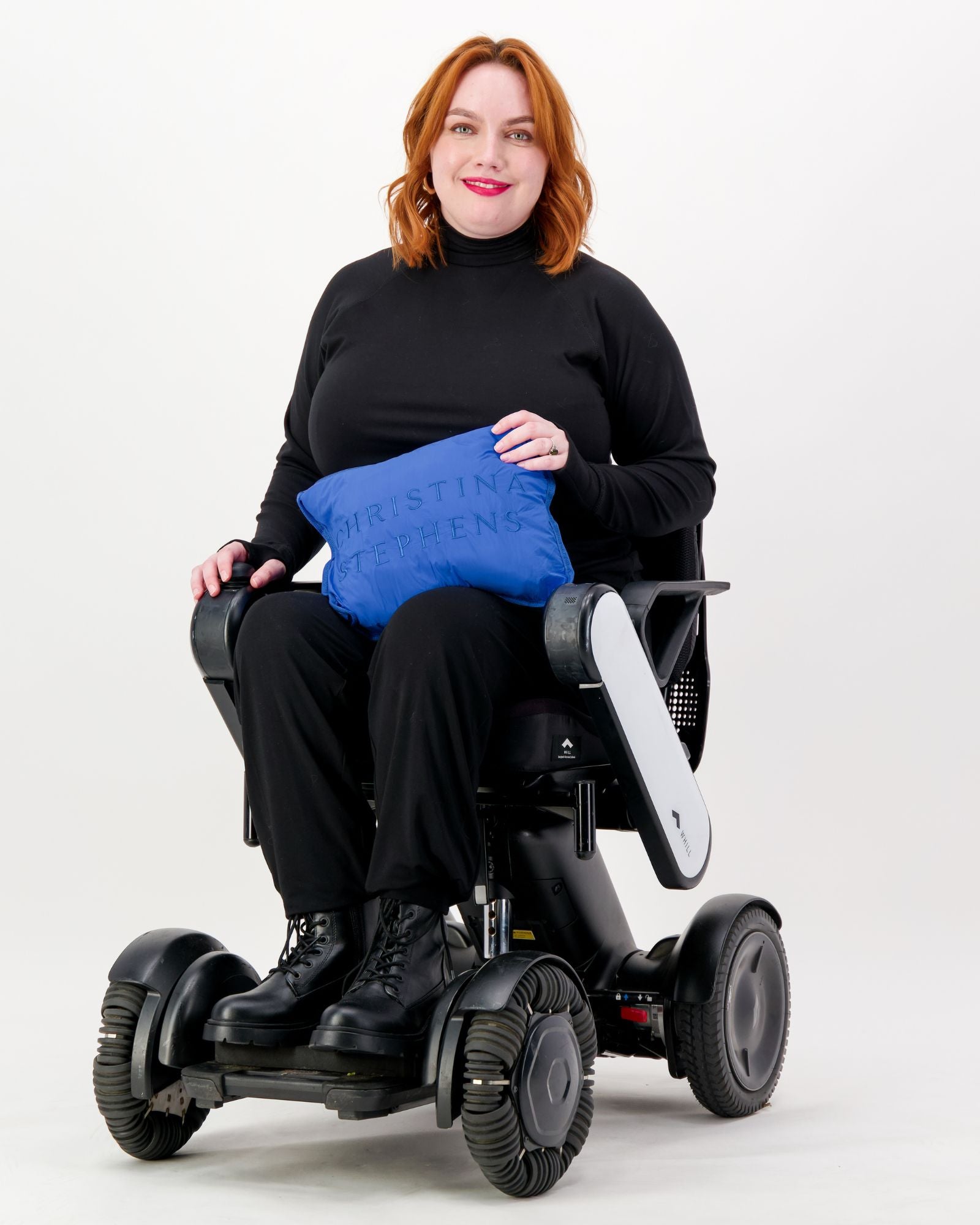 Elly Universal Puffer Jacket demonstrated on a power wheelchair user wearing black attire, holding a folded blue version of the jacket that converts to a travel pillow. The adaptive jacket showcases its versatility and practical design against a white background.