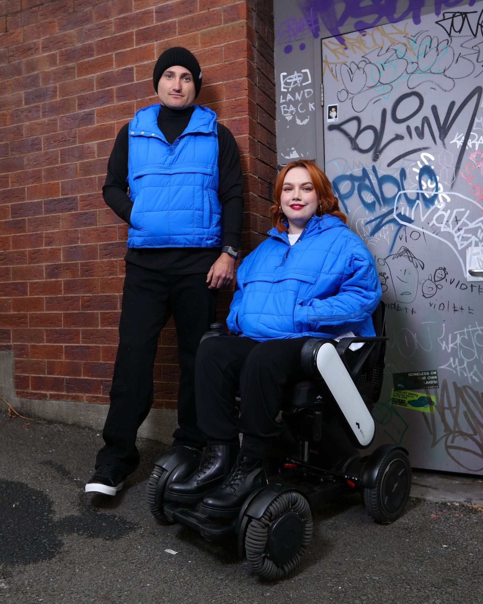 Elly Universal Puffer Jacket shown in vibrant blue, displayed against a brick wall with graffiti. Models demonstrate versatile wear for both standing and seated positions, highlighting the adaptive design features and wheelchair-friendly functionality.