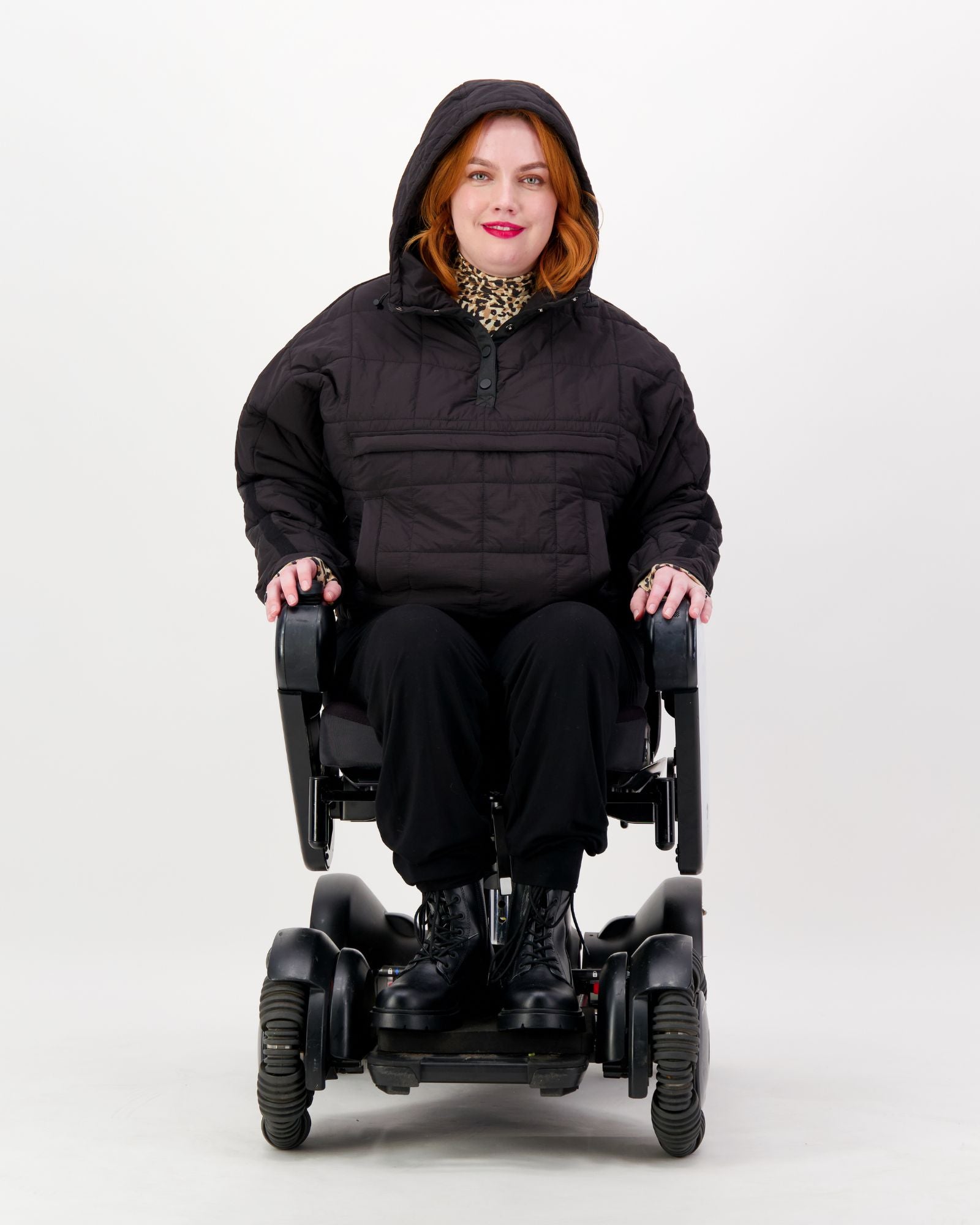 Elly Universal Puffer Jacket shown on a wheelchair user against a white background, featuring a black hooded design with pull-over style. The adaptive jacket demonstrates its wheelchair-friendly fit while paired with black pants and boots, highlighting its practical and stylish design.