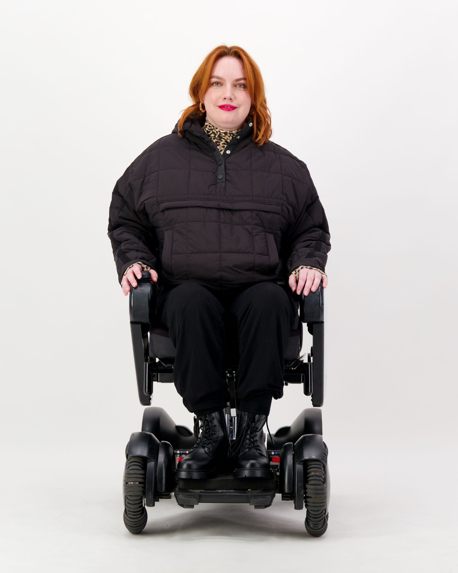 Elly Universal Puffer Jacket shown on a wheelchair user, featuring a black quilted design with front pocket. The adaptive jacket demonstrates its comfortable fit in a seated position, paired with black pants and boots against a white background.