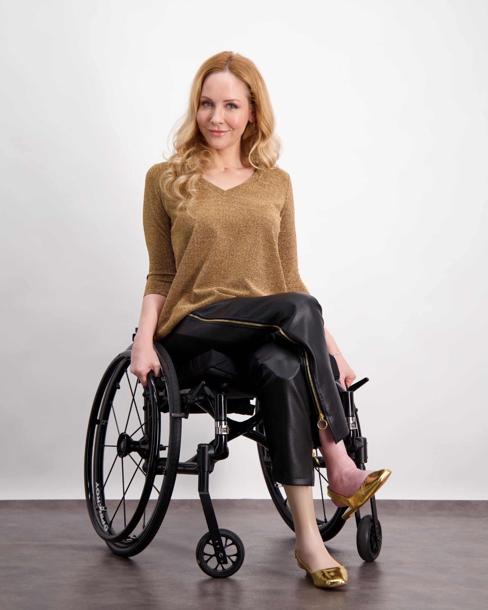 Model wearing Suzie Q Seated Pants in sleek black pleather, paired with a gold V-neck sweater and metallic flats, showcasing adaptive design features while seated in a wheelchair against a light background.