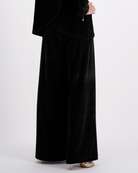 Jessie Wide Leg Velvet Pants shown in luxurious black velvet fabric, featuring an elegant wide-leg silhouette and side pull tabs, styled with metallic heels against a white background.