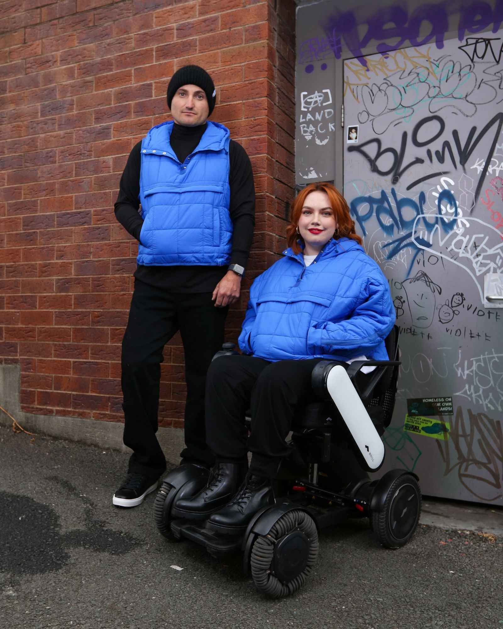 Matt Gender Neutral Puffer Vest shown in vibrant blue, modeled against a brick wall with graffiti. The versatile vest features a comfortable design that can be worn while seated or standing, demonstrating its adaptive functionality and stylish urban aesthetic.