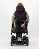 Matt Gender Neutral Puffer Vest shown from back view on person using power wheelchair. Black quilted vest paired with leopard print sleeves, demonstrating adaptable design and wheelchair-friendly fit. Vest features packable design and adaptive elements for comfort while seated.