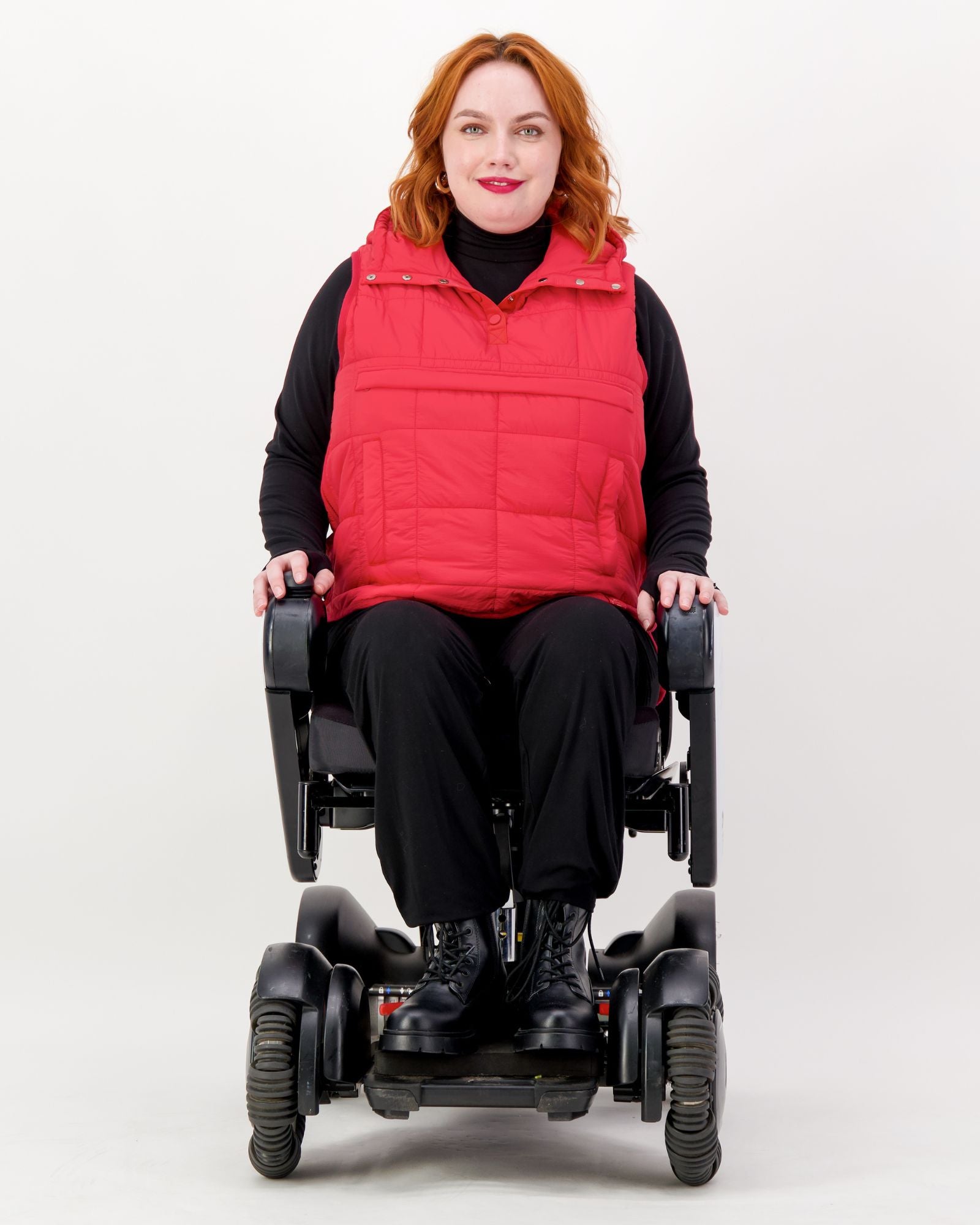 Matt Gender Neutral Puffer Vest shown on a model in a wheelchair against a white background. The bright red vest features a high collar and quilted design, worn over a black turtleneck with black pants and boots, demonstrating adaptive comfort and mobility.