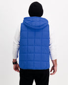 Matt Gender Neutral Puffer Vest shown from back view in vibrant royal blue, featuring quilted design, hooded collar, and sleeveless style. Displayed over white long-sleeve shirt with black beanie, demonstrating its layering versatility and adaptive design.