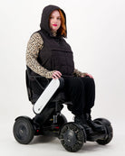 Matt Gender Neutral Puffer Vest shown on a wheelchair user wearing a leopard print top. Black hooded vest features adaptive design for wheelchair comfort. Model seated in a modern white electric wheelchair against white background.