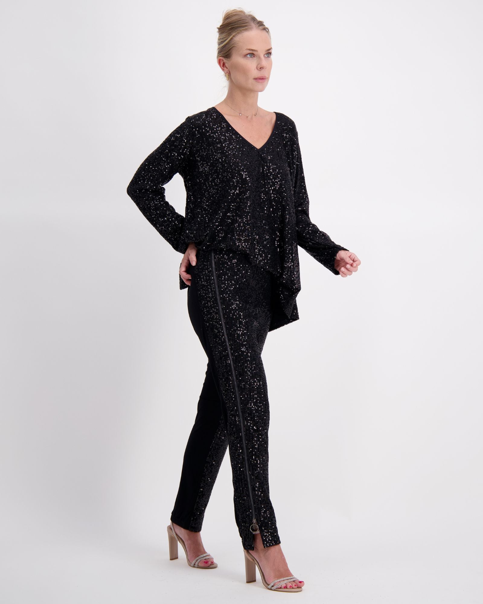 Suzie Q Seated Pants Sequins shown in elegant black sequin fabric. Model wearing matching v-neck tunic top with long sleeves, creating a stunning evening ensemble. Adaptive pants feature seamless design and accommodating fit, styled with nude heels against white background.