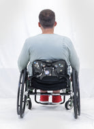 Back view of Men's Long Sleeve Leaf Back T-Shirt shown on a person in a wheelchair, wearing a light grey shirt with red shoes, demonstrating the comfortable fit and casual style against a white background.