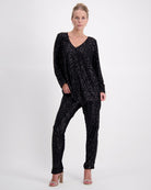 Tara V Neck Leaf Back Top shown in black metallic glitter fabric with full-length sleeves and V-neckline, styled as a complete outfit with matching sequined pants and neutral heels against a white background