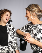 Lilli Leaf Back T-Shirt shown in black and white floral print bamboo fabric, demonstrating joyful interaction between two people wearing matching patterns, highlighting the comfort and style of this sensory-friendly adaptive clothing design against a gray studio background.