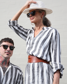 Sapphire Magnetic Shirt Dress styled with a wide-brim white hat and brown leather belt, showcasing vertical grey and white stripes in a casual outdoor setting against a white wall. The linen dress features long sleeves and a relaxed silhouette perfect for easy dressing.