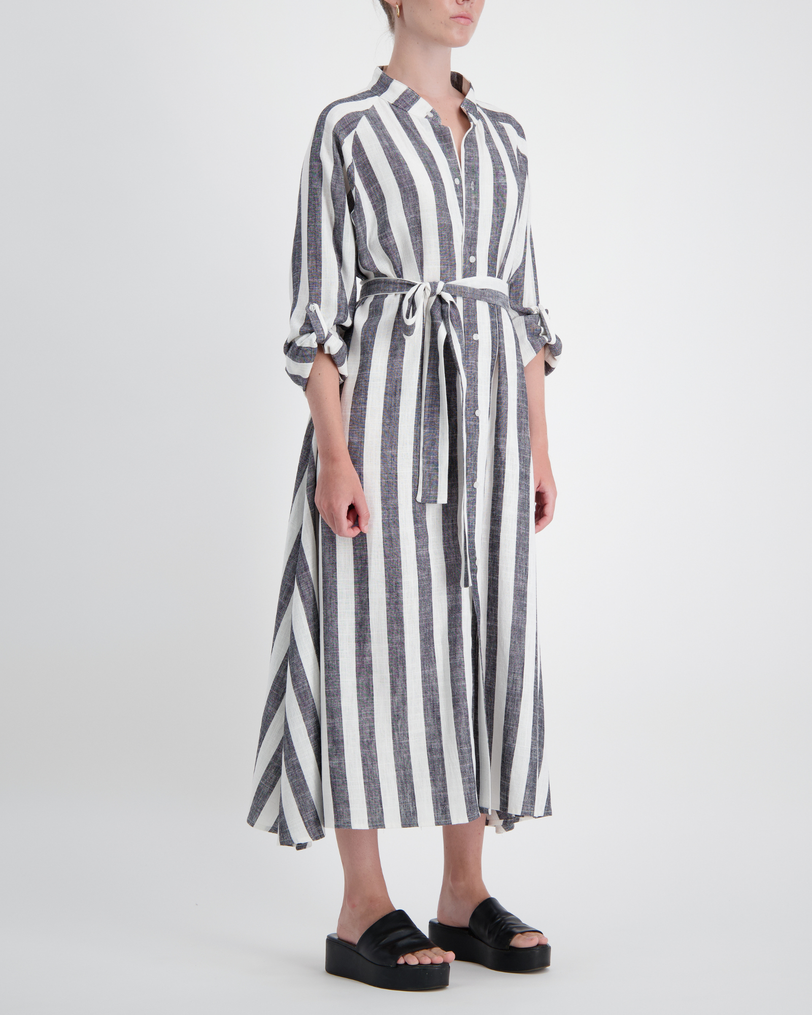 Sapphire Magnetic Shirt Dress in bold grey and white vertical stripes, featuring three-quarter rolled sleeves, collared neckline, and a waist-defining belt tie. The linen dress flows elegantly to midi length, shown paired with black platform sandals against a light background.