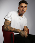 Nick A-Line Tee showcased in white SpillFree fabric, featuring a classic V-neck design. Model seated in burgundy chair displaying the adaptive tee's comfortable fit and modern silhouette against a gray studio background, highlighting detailed tattoo artistry.