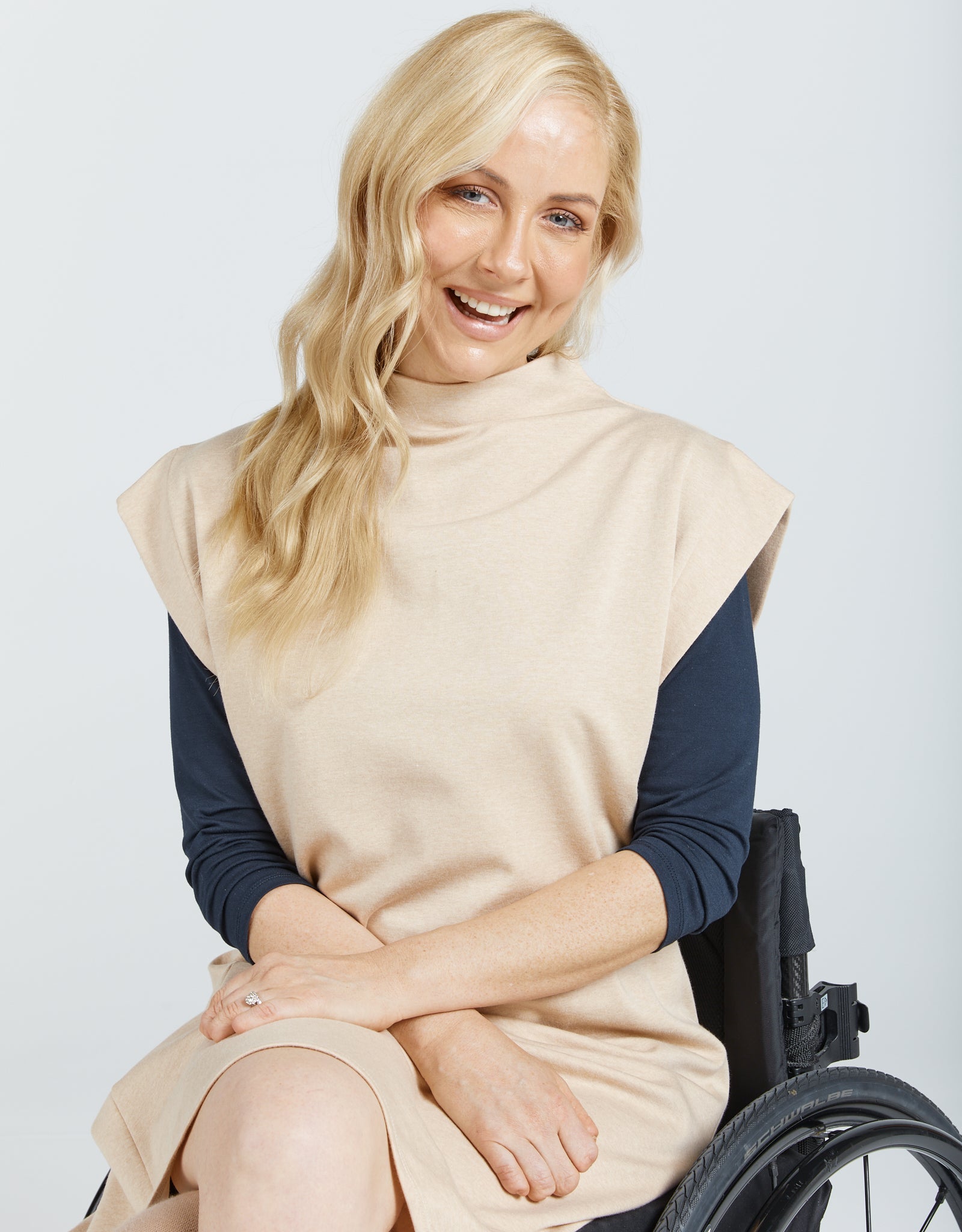 Leaf Back Dress shown on a model in wheelchair, featuring a beige sleeveless design with high neckline and navy long sleeves underneath. Natural cotton fabric in neutral tone against white background displays modern, adaptive fashion styling.