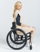 Leaf Back Dress shown in beige cotton fabric on model using wheelchair, featuring unique leaf-shaped back design, high neckline, and knee-length cut. Navy long sleeves contrast with natural undyed dress body, photographed against white background.