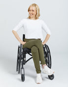 Seated Leggings shown on a wheelchair user wearing a white top, demonstrating the olive green leggings' comfortable fit and easy-wear design. The leggings feature a streamlined silhouette paired with white sneakers against a light background.