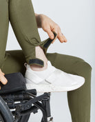 Seated Legging for SP Catheter shown in olive green color, with close-up detail of adjustable ankle opening being demonstrated with white sneaker. Special design features reinforced access points and functionality for medical devices.