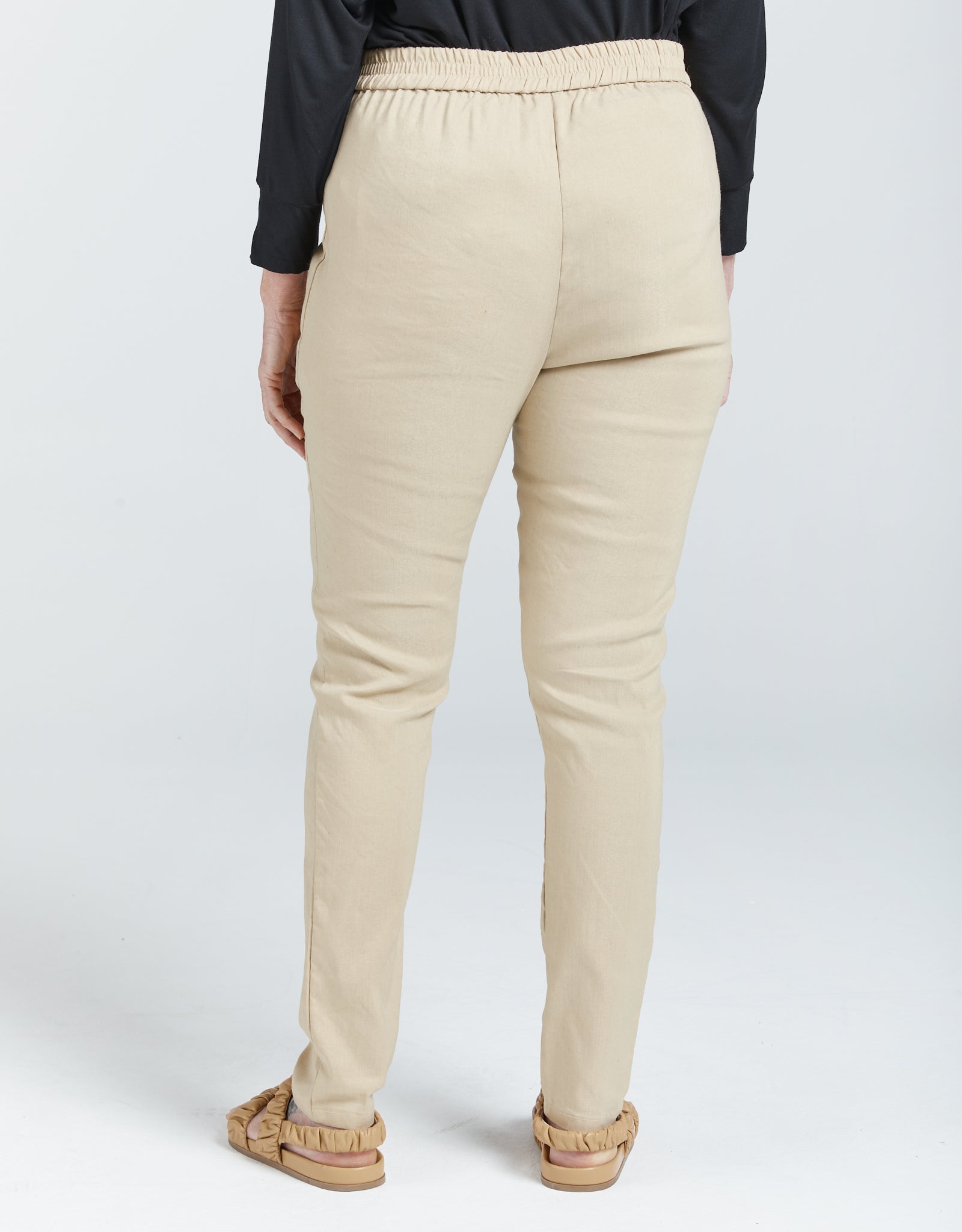 Seated Stretch Pant shown from back view in beige color with elastic waistband, straight-leg design, and full-length fit. Model wearing black top and tan sandals against white background, demonstrating comfortable, tailored silhouette ideal for wheelchair users.