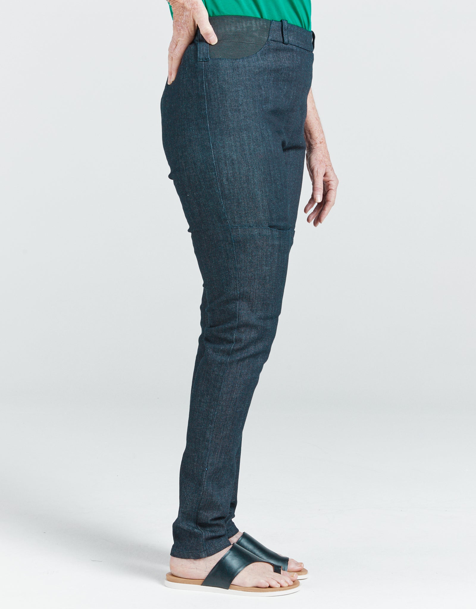 Seated Stretch Pant shown in dark denim side view, featuring sleek fitted design with high-rise back waistband, paired with green top and black sandals against light background