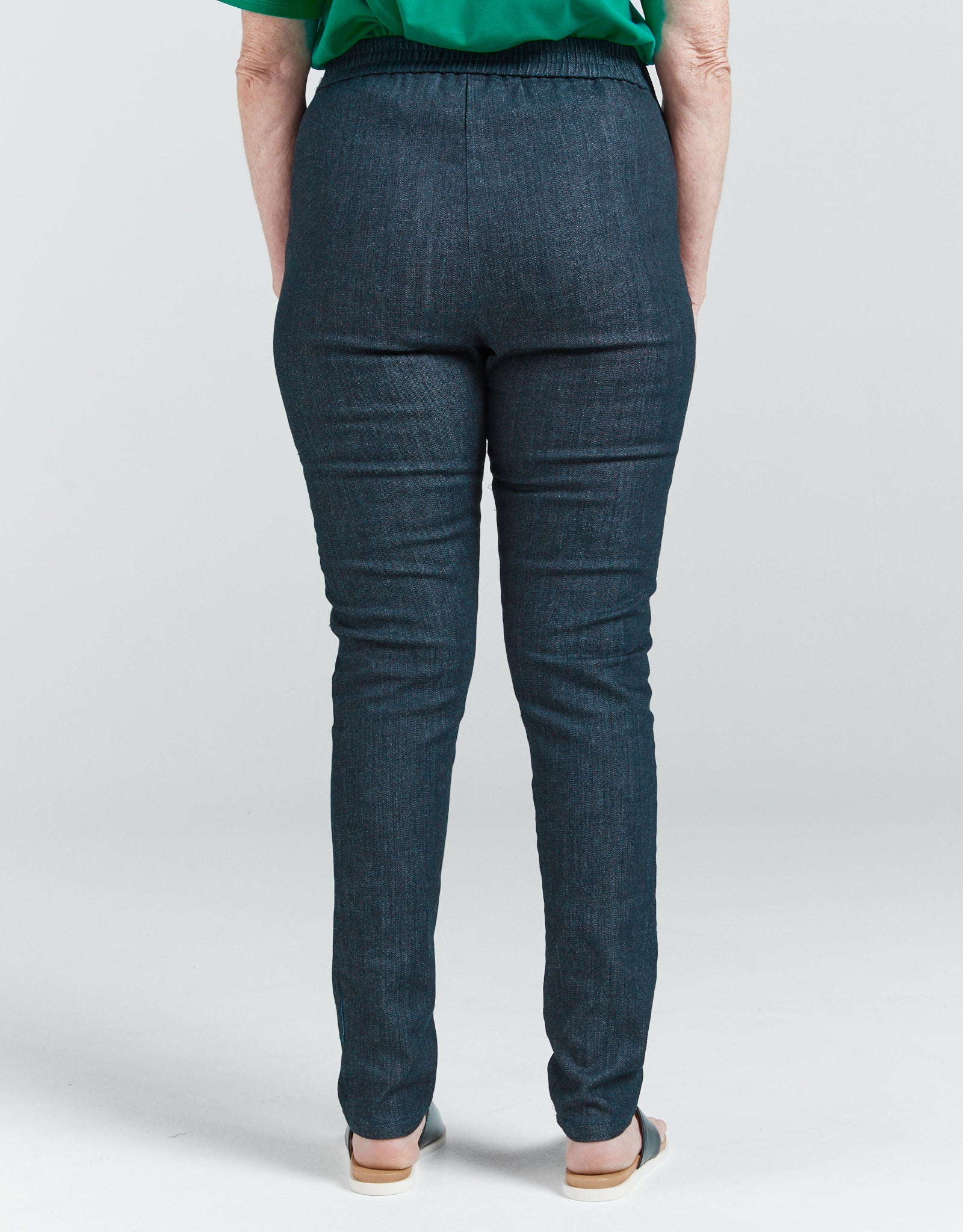 Seated Stretch Pant shown from back view, featuring dark denim fabric with elastic waistband and sleek tailored fit. Model wearing emerald green top, demonstrating the pants' full coverage design and smooth back panel construction.