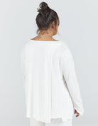 Essensual PJ Top shown from back view featuring a relaxed white long-sleeve design with boat neckline, loose-fitting silhouette, and soft draping fabric. Model wearing casual messy bun hairstyle against light gray background.