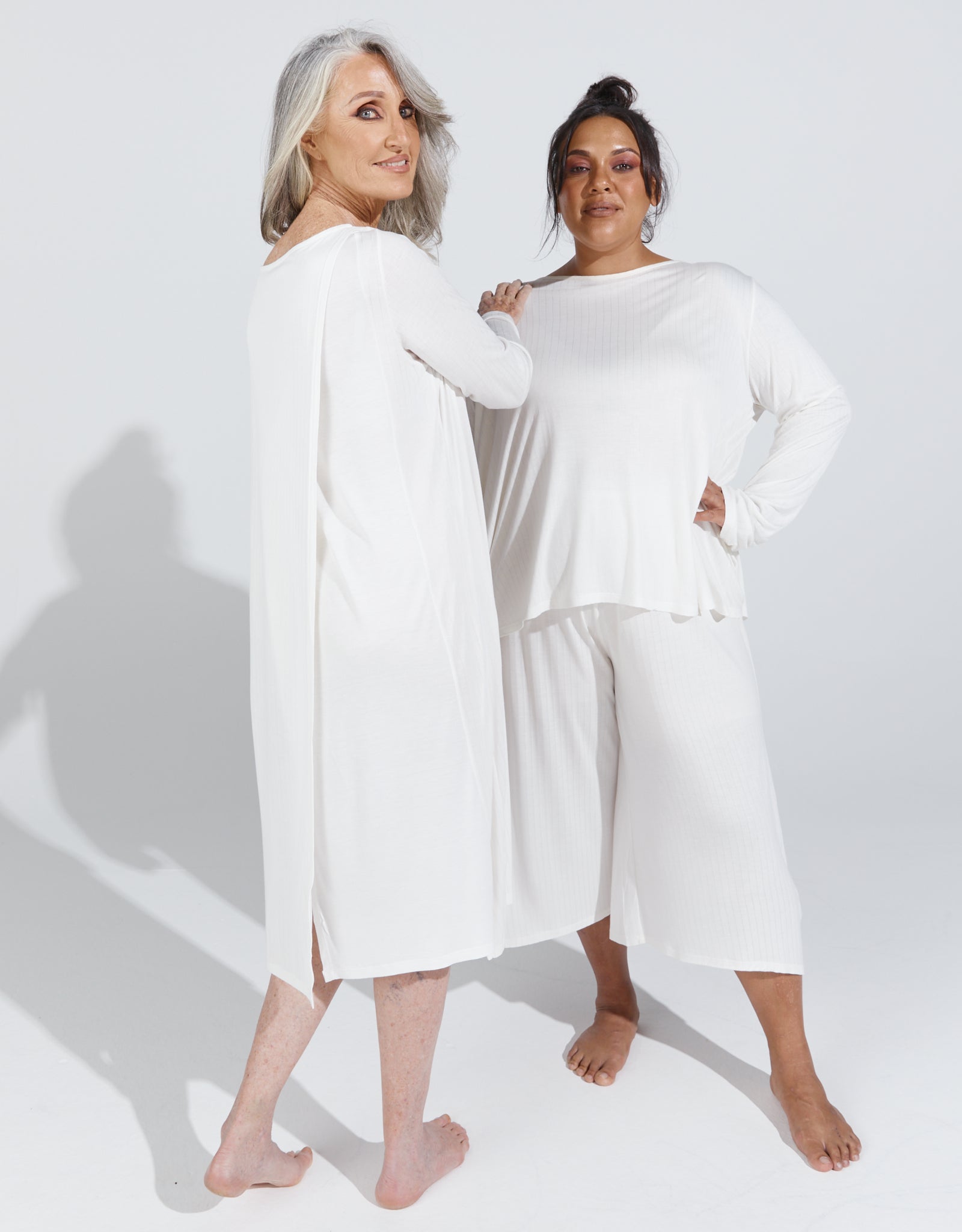 Two models wearing the Essensual PJ Dress in pure white, showcasing the long-sleeved, relaxed-fit design with mid-calf length. The sleepwear features comfortable, flowing fabric ideal for restful nights.