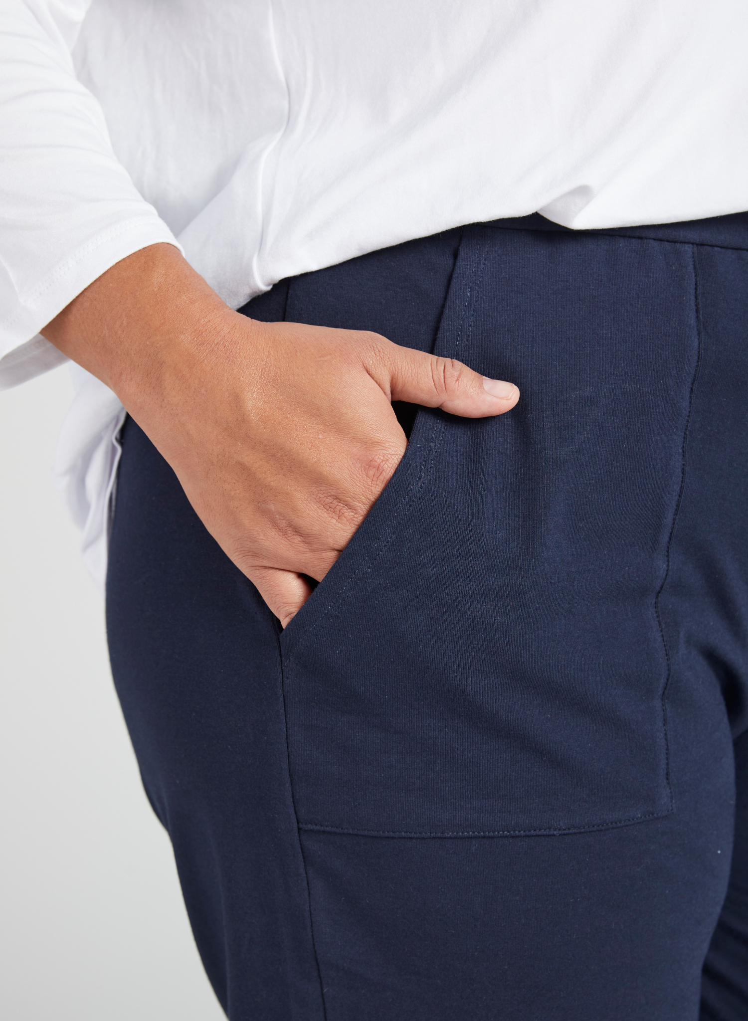 Tapered Track Pants with Adjustable Cuff shown in navy blue, featuring a close-up view of the side pocket detail with a hand demonstrating the pocket placement. White top visible in background highlighting the mid-rise waistband design.