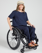 Model wearing Short Sleeve Leaf Back T-Shirt in navy blue, styled with beige cap and orange sandals, seated in a modern wheelchair against light grey background, demonstrating adaptive fashion and inclusive design.