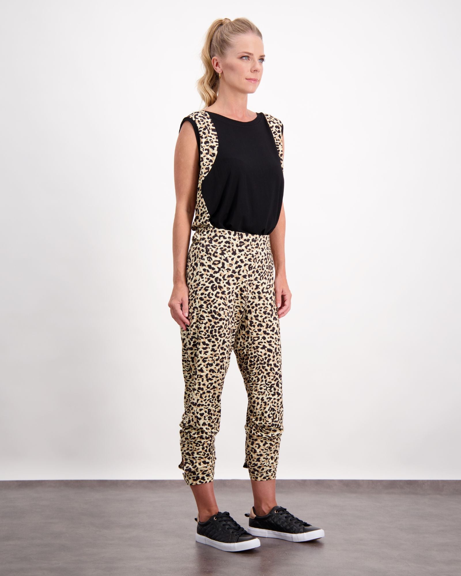 Lauren Sleeveless Physio Top shown in black, paired with leopard print pants, creating a stylish athletic ensemble. The sleeveless design features a classic round neckline and relaxed fit, perfect for exercise or casual wear. Model stands against white background wearing black sneakers.