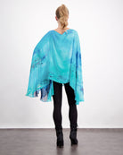 Oceania Kaftan shown from behind, featuring flowing aqua blue silk with darker blue watercolor patterns, draped elegantly over black leggings. The luxurious fabric creates a dramatic, wing-like silhouette against a white background.