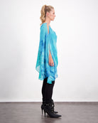 Oceania Kaftan shown in side profile, featuring flowing aqua blue silk with watercolor-like patterns. The luxurious kaftan drapes elegantly over dark pants, paired with black stiletto boots. The ethereal design showcases a versatile silhouette perfect for evening wear and special occasions.