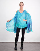 Oceania Kaftan featuring flowing silk fabric in vibrant aqua and deep blue watercolor design, with elegant cold-shoulder cut and dramatic cape-like draping, paired with black leggings and boots against a white background