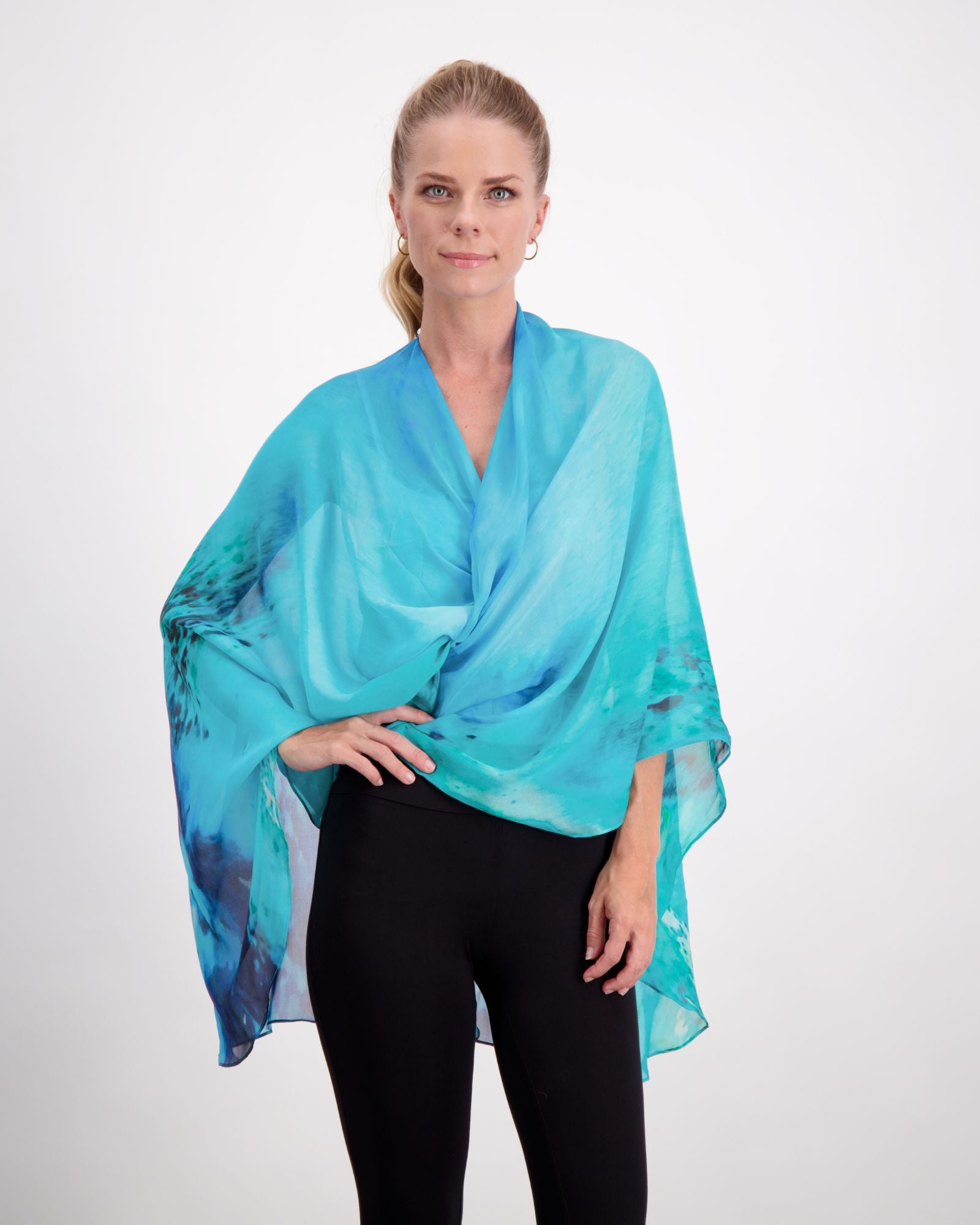 Oceania Kaftan displayed as an elegant aqua blue silk wrap with navy abstract patterns draped stylishly over black pants, showcasing its versatile styling potential and luxurious flowing fabric against a white background