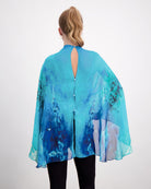 Back view of Oceania Kaftan featuring flowing silk fabric in vibrant aqua and deep blue watercolor patterns, with elegant keyhole button closure and dramatic cape-like silhouette against white background. Black pants complete the ensemble.