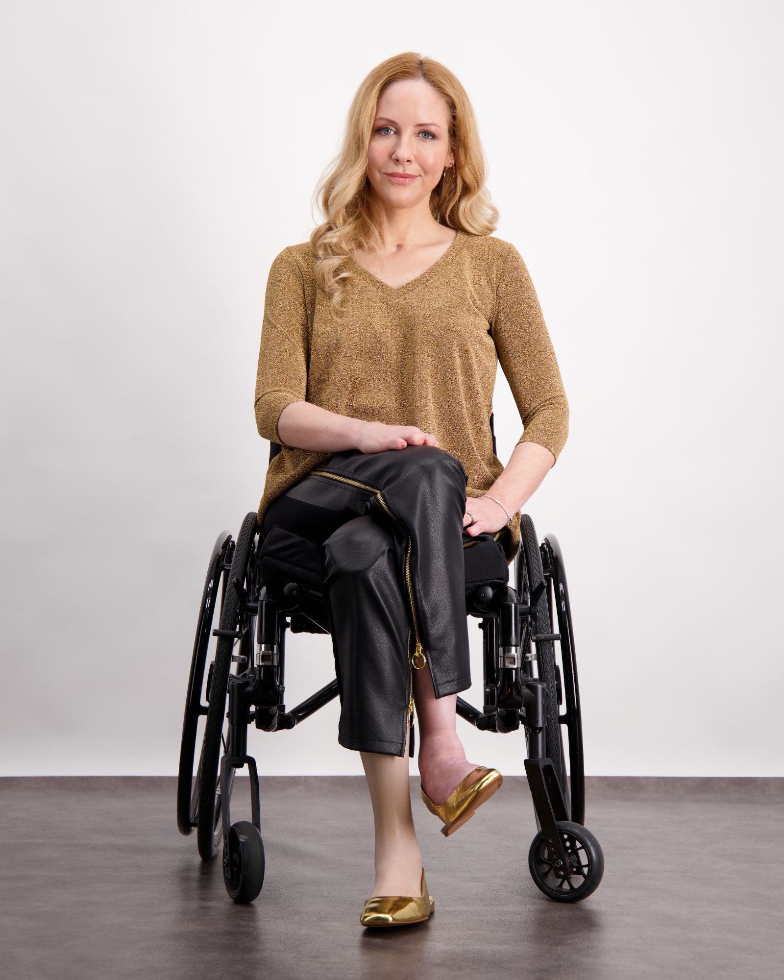 Suzie Q Seated Pants shown on model in wheelchair, featuring black pleather material with gold zippers, paired with metallic gold v-neck top and matching flats. Adaptive design visible with spacious fit and accessible zipper details against white studio background.