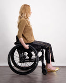 Suzie Q Seated Pants shown on a model in a wheelchair, featuring a black adaptive design with gold accents, paired with a brown sweater and metallic flats. Side view demonstrates seamless fit and wheelchair-friendly construction.