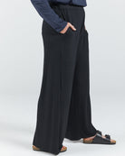 Jessie Wide Leg Pant shown from hip to ankle, featuring a flowing black silhouette with side pockets and relaxed fit. Model wears dark sandals. Navy blue top partially visible. Photographed against light grey background, highlighting the pants' elegant drape and comfort-focused design.