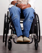 Stephen Seated Jeans shown on a person in a wheelchair, featuring classic blue denim with side pockets. The adaptive jeans are paired with a white top and beige shoes, demonstrating comfortable seated wear and accessibility-focused design.