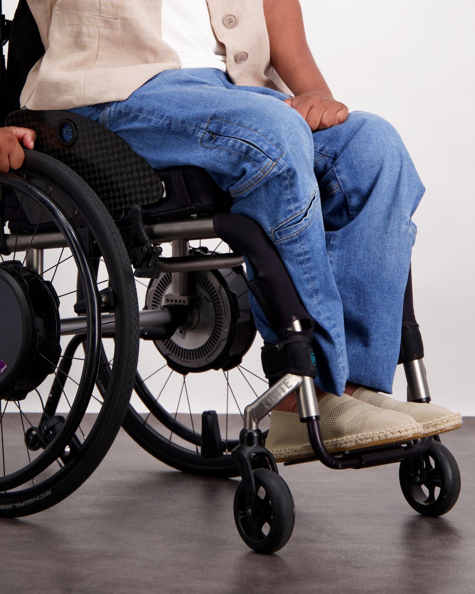 Stephen Seated Jeans shown on a wheelchair user, featuring comfortable blue denim designed for seated wear. Detail view highlighting adaptive features, paired with a beige vest and casual footwear. Wheelchair visible in frame demonstrating practical accessibility features.