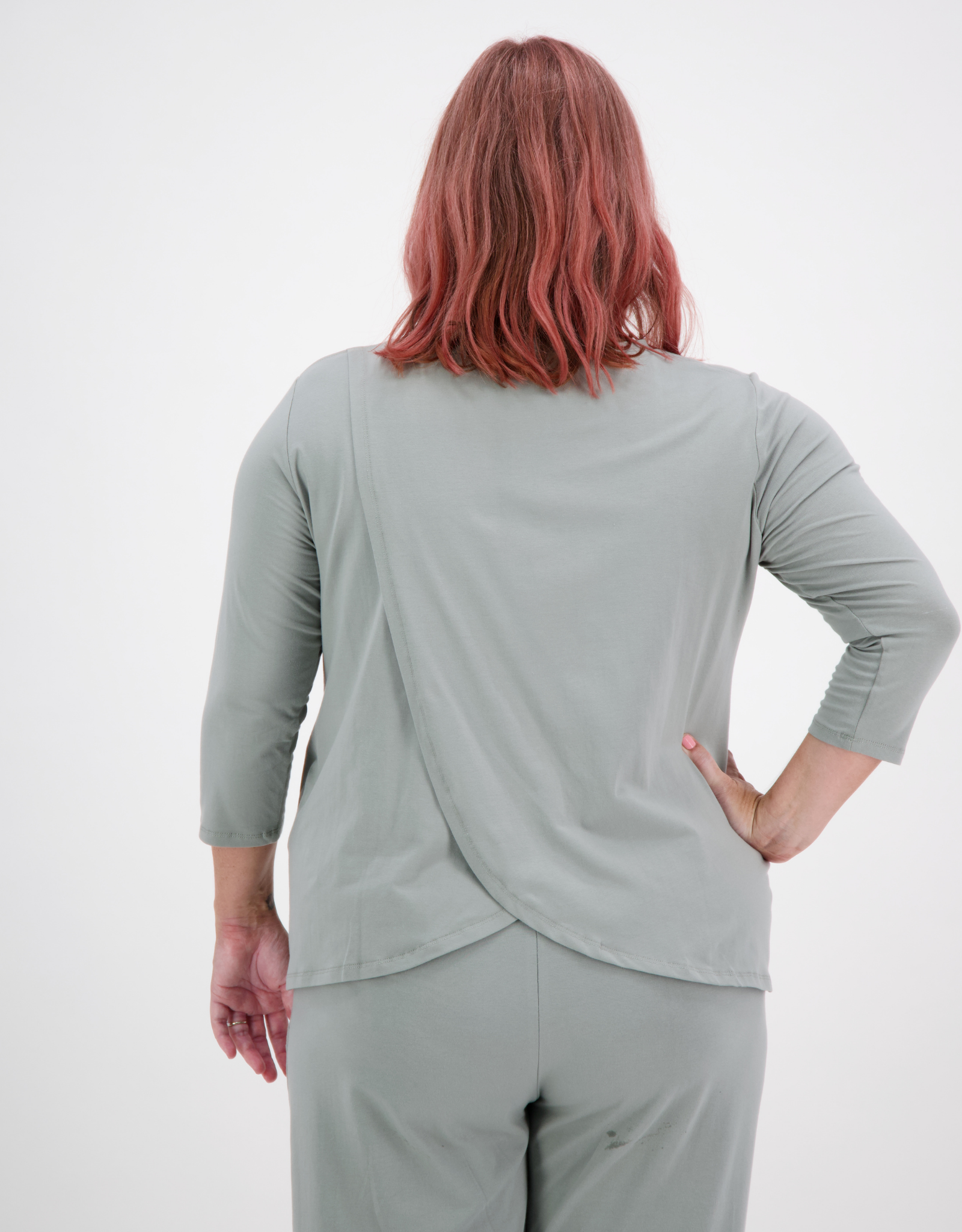 Back view of 3/4 Sleeve Leaf Back T-Shirt in soft sage green, showcasing distinctive overlapping leaf design at lower back, styled with matching pants. Rose-colored hair model demonstrates adaptive stretch and elegant drape of the garment.