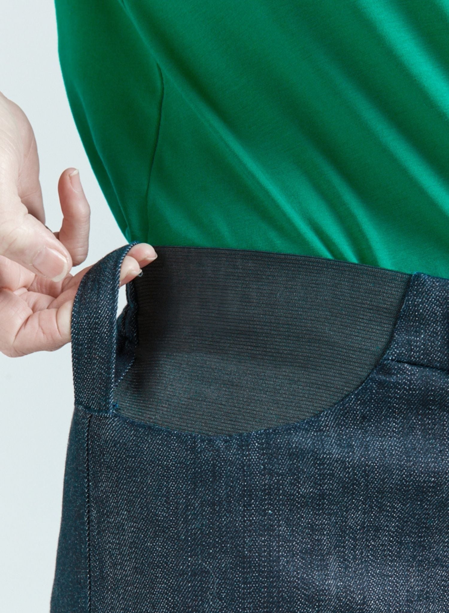 Seated Stretch Pant featuring an elastic waistband detail in dark denim, shown against an emerald green top. Close-up view highlighting the adaptable stretch panel design that prevents riding up while seated, demonstrating the pants' functional yet stylish construction.