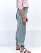 Tapered Track Pants with Adjustable Cuff shown in sage green color, styled with a pink top. Side view displaying the relaxed fit, elastic waistband, and tapered leg design with adjustable cuffs at ankles, paired with casual sandals.